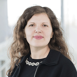 A smiling woman wearing a black blazer stands in front of a window and looks at the camera.